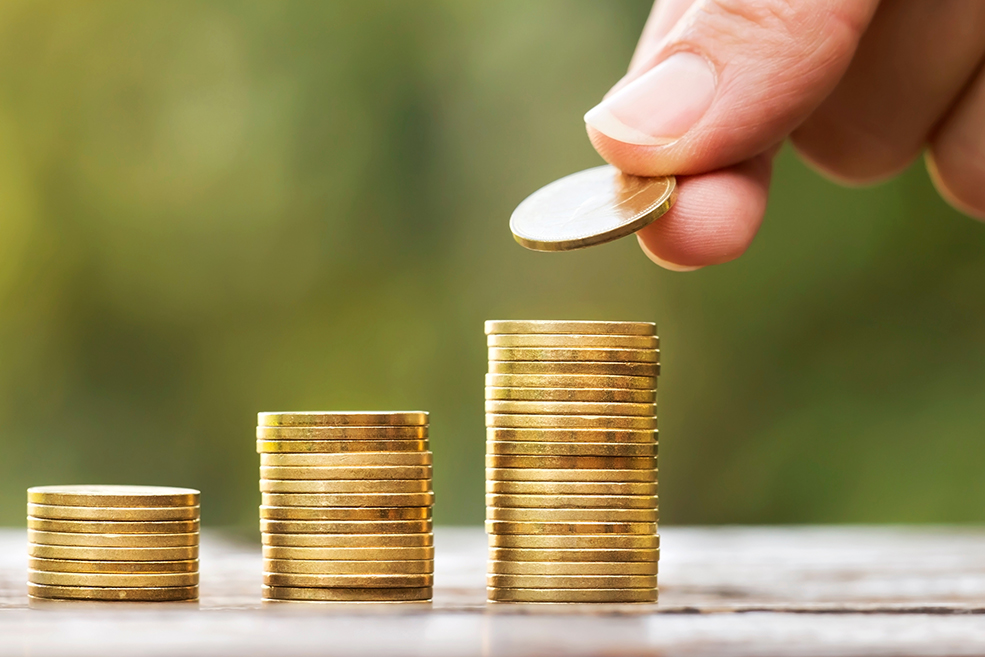 Three stacks of gold coins of increasing size with a hand adding a coin to the third stack.